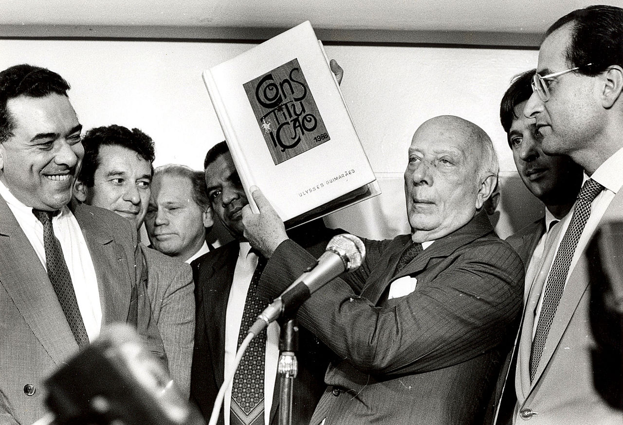 A group of men stand behind a micrphone. One holds up a book with the word "Constitution" in portuguese on the front cover.